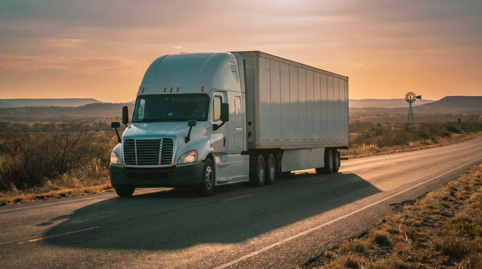 A white semi truck alone on an open two-lane road at sunset, a windmill on the plain behind it.