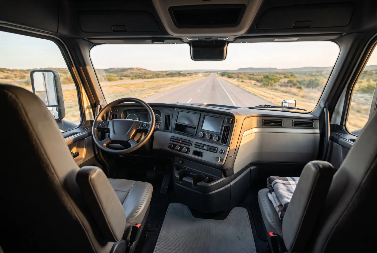 Inside a truck cab, seen from behind the seats: the steering wheel and dashboard, and the windshield looking out on an open highway at golden hour.