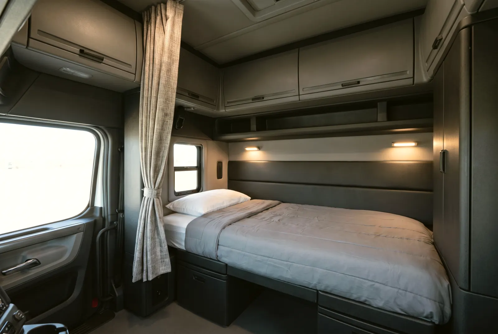 The sleeper berth of a truck cab: a made-up bunk with a grey blanket and a white pillow, cabinets overhead, reading lights and a small window.